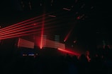 Red laser lights create dynamic lines across a dark club setting, illuminating a crowd of people. The atmosphere is energetic and vibrant with various angles of beams cutting through the dimly lit room.