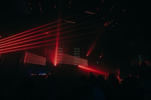 Red laser lights create dynamic lines across a dark club setting, illuminating a crowd of people. The atmosphere is energetic and vibrant with various angles of beams cutting through the dimly lit room.