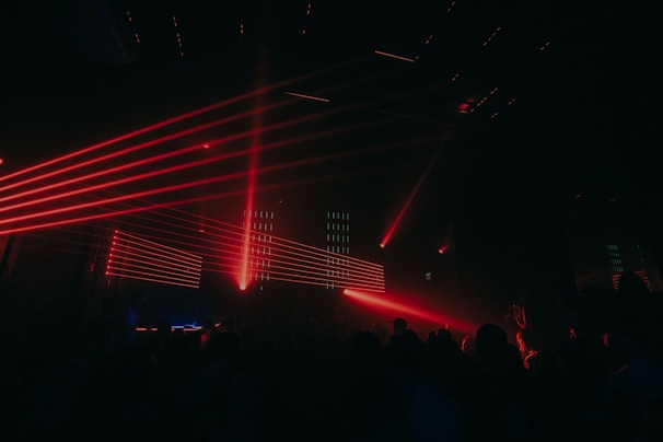 Red laser lights create dynamic lines across a dark club setting, illuminating a crowd of people. The atmosphere is energetic and vibrant with various angles of beams cutting through the dimly lit room.