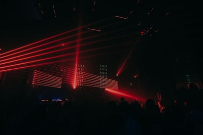 Red laser lights create dynamic lines across a dark club setting, illuminating a crowd of people. The atmosphere is energetic and vibrant with various angles of beams cutting through the dimly lit room.