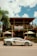 A sleek silver sports car is parked in front of a rustic wooden restaurant called Buena Vibra. The building has a wooden balcony, and there are outdoor umbrellas shading tables. A large palm tree is visible to the left, and the sky is partly cloudy.