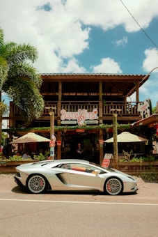 A sleek silver sports car is parked in front of a rustic wooden restaurant called Buena Vibra. The building has a wooden balcony, and there are outdoor umbrellas shading tables. A large palm tree is visible to the left, and the sky is partly cloudy.