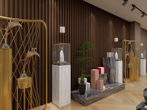 A stylish and modern interior with a deep brown, slatted wooden wall. Several decorative elements, such as golden structures with animal antler decorations, are placed in the foreground. White pedestals topped with glass cases hold uniquely designed bottles. A small potted plant adds greenery, and there are geometric stone sculptures in varied colors. The subtle lighting creates a warm and inviting atmosphere.