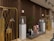A stylish and modern interior with a deep brown, slatted wooden wall. Several decorative elements, such as golden structures with animal antler decorations, are placed in the foreground. White pedestals topped with glass cases hold uniquely designed bottles. A small potted plant adds greenery, and there are geometric stone sculptures in varied colors. The subtle lighting creates a warm and inviting atmosphere.