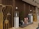 A stylish and modern interior with a deep brown, slatted wooden wall. Several decorative elements, such as golden structures with animal antler decorations, are placed in the foreground. White pedestals topped with glass cases hold uniquely designed bottles. A small potted plant adds greenery, and there are geometric stone sculptures in varied colors. The subtle lighting creates a warm and inviting atmosphere.