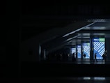In a low-light environment, silhouetted figures walk in front of brightly lit digital displays featuring abstract geometric patterns and text. The setting appears to be a modern, underground or indoor transit area with high ceilings and structural beams.