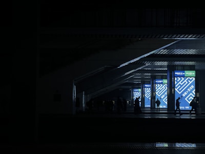 In a low-light environment, silhouetted figures walk in front of brightly lit digital displays featuring abstract geometric patterns and text. The setting appears to be a modern, underground or indoor transit area with high ceilings and structural beams.