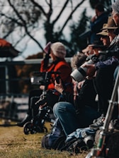 A group of people is gathered outdoors, possibly at a sports or public event. Some individuals are sitting or standing with cameras, wearing casual attire, including jackets and hats. A black dog is also present near one seated person.