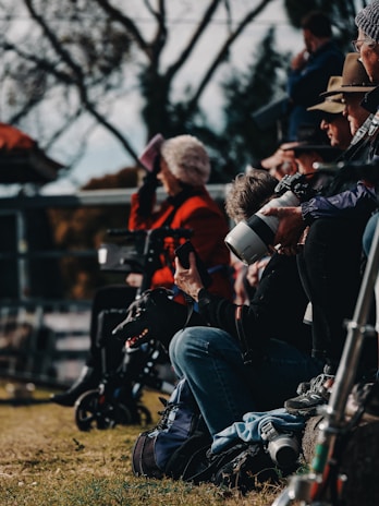 A group of people is gathered outdoors, possibly at a sports or public event. Some individuals are sitting or standing with cameras, wearing casual attire, including jackets and hats. A black dog is also present near one seated person.