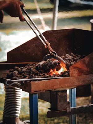 A person uses metal tongs to manipulate hot coals or embers in a forge. The coals are glowing with intense heat, suggesting an active blacksmithing or metalworking process. The surrounding area appears industrial, with metal surfaces and equipment visible.