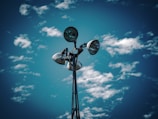 A tall metal light pole with multiple spotlights is set against a vibrant blue sky speckled with scattered white clouds. The lights are positioned at various angles, catching the daylight.