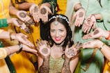 A woman, elegantly dressed, is surrounded by several individuals showcasing intricate henna designs on their hands. The woman's vibrant smile and elaborate jewelry accentuate the celebratory atmosphere.