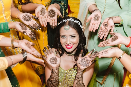 A woman, elegantly dressed, is surrounded by several individuals showcasing intricate henna designs on their hands. The woman's vibrant smile and elaborate jewelry accentuate the celebratory atmosphere.