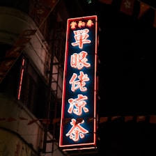 A neon sign with Chinese characters glows brightly against a dark background, mounted on a building. The sign is illuminated with a combination of red and blue lights. Nearby are small flags featuring a floral design and a portion of a star, contributing to a festive or traditional atmosphere.