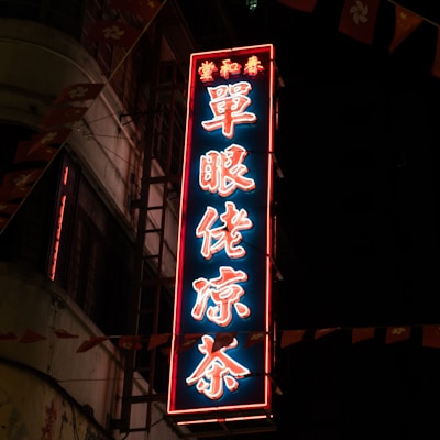 A neon sign with Chinese characters glows brightly against a dark background, mounted on a building. The sign is illuminated with a combination of red and blue lights. Nearby are small flags featuring a floral design and a portion of a star, contributing to a festive or traditional atmosphere.