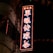 A neon sign with Chinese characters glows brightly against a dark background, mounted on a building. The sign is illuminated with a combination of red and blue lights. Nearby are small flags featuring a floral design and a portion of a star, contributing to a festive or traditional atmosphere.