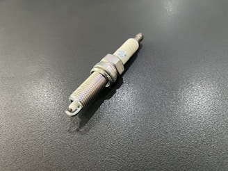 A single spark plug is placed on a dark, textured surface. The metallic body and ceramic insulator are visible, showcasing its typical design.