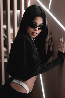 A person with long black hair is leaning against a wall with illuminated lines. They are wearing dark sunglasses, a black cropped top, and have visible metallic details on their pants. The setting has a modern aesthetic with wooden and illuminated elements.