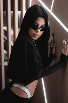 A person with long black hair is leaning against a wall with illuminated lines. They are wearing dark sunglasses, a black cropped top, and have visible metallic details on their pants. The setting has a modern aesthetic with wooden and illuminated elements.