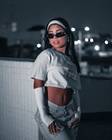 A stylish person is posing against a tiled wall in a dimly lit urban setting. They are wearing sunglasses, a white crop top, high-waisted pants, and long white sleeves, exuding a modern and confident aesthetic. The background features bokeh lights that enhance the nighttime atmosphere.