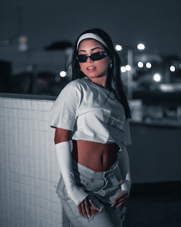 A stylish person is posing against a tiled wall in a dimly lit urban setting. They are wearing sunglasses, a white crop top, high-waisted pants, and long white sleeves, exuding a modern and confident aesthetic. The background features bokeh lights that enhance the nighttime atmosphere.