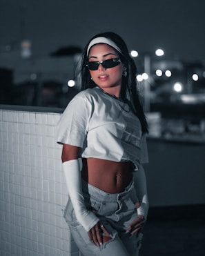 A stylish person is posing against a tiled wall in a dimly lit urban setting. They are wearing sunglasses, a white crop top, high-waisted pants, and long white sleeves, exuding a modern and confident aesthetic. The background features bokeh lights that enhance the nighttime atmosphere.