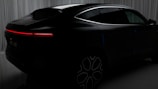 A sleek black car with a futuristic design is set against a backdrop of curtains. The vehicle features a seamless, aerodynamic shape with a prominent red LED tail light stretching horizontally across the rear. The wheels have a modern, intricate pattern, adding to the overall sophisticated appearance of the car.