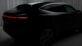 A sleek black car with a futuristic design is set against a backdrop of curtains. The vehicle features a seamless, aerodynamic shape with a prominent red LED tail light stretching horizontally across the rear. The wheels have a modern, intricate pattern, adding to the overall sophisticated appearance of the car.