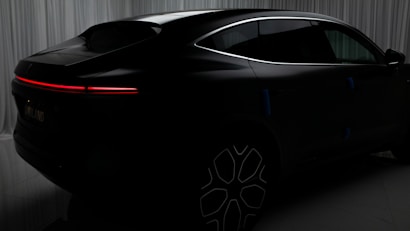 A sleek black car with a futuristic design is set against a backdrop of curtains. The vehicle features a seamless, aerodynamic shape with a prominent red LED tail light stretching horizontally across the rear. The wheels have a modern, intricate pattern, adding to the overall sophisticated appearance of the car.