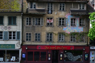 An old building with worn-out facades featuring multiple windows, some of which have open shutters. The structure shows signs of decay and is partially covered with ivy. A pirate flag hangs from one window and there is colorful graffiti on the wall. The ground floor houses a shop with a red sign that reads 'The Bruch Brothers.'