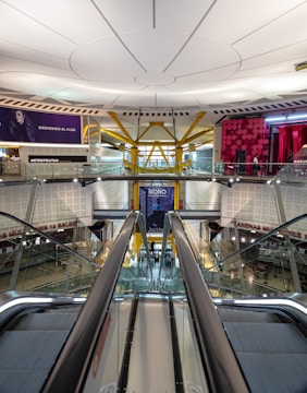 A modern indoor architectural space featuring an escalator leading to an upper level. The ceiling is designed with circular patterns and there are colorful banners and advertisements displayed around. Glass railings and bright lighting add to the contemporary atmosphere.