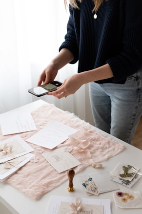 A person wearing a dark sweater and jeans is taking a photo with a smartphone. The table is covered with pink fabric, and several elegant paper items including invitations, envelopes, and a stamp are arranged neatly. Also visible are a wax seal stamp and some decorative dried flowers.