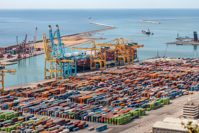 Numerous colorful shipping containers neatly arranged in a large port area near the ocean. Several large cranes are present, indicating an active shipping or logistics hub. The vast expanse of the sea in the background highlights the scale of the port.