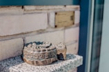 A metallic sculpture with intricate carvings resembling a crown or wreath sits on a stone or concrete ledge. The background features a light-colored brick wall with a bronze plaque.
