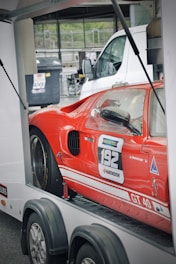 A red sports car with the number 192 on the side sits on a trailer. The car has sponsorship decals including Ravenol and Hankook. White detailing and the label GT 40 are visible on the lower side panel. The background shows a parking area with other vehicles and a large dumpster.