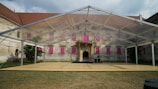A transparent tent structure is set up on a grassy area in front of an old, rustic building with visible wear and tear. The building has numerous windows covered with bright pink panels, and there are small trees or bushes on both sides. The sky above is partly cloudy.