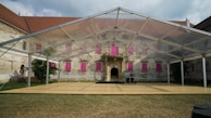 A transparent tent structure is set up on a grassy area in front of an old, rustic building with visible wear and tear. The building has numerous windows covered with bright pink panels, and there are small trees or bushes on both sides. The sky above is partly cloudy.