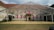 A transparent tent structure is set up on a grassy area in front of an old, rustic building with visible wear and tear. The building has numerous windows covered with bright pink panels, and there are small trees or bushes on both sides. The sky above is partly cloudy.