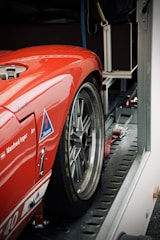 A red sports car is securely fastened inside a trailer. The focus is on the rear section, highlighting the sleek metallic wheel and tire. Visible branding and sponsor logos add a racing aesthetic to the scene.