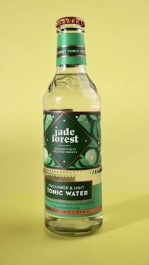 A clear glass bottle of tonic water with a green label featuring leaf and cucumber slice illustrations. The label displays the brand name 'jade forest' and description of the flavor, cucumber and mint. The bottle has a copper-colored cap and a vibrant green background.