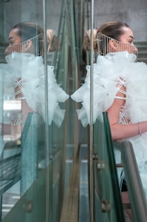 A person is standing beside a glass railing, creating a mirrored reflection. They are wearing a white, ruffled, and elaborate outfit with detailed embellishments on the face and a hair accessory. The setting appears to be indoors, likely in a modern architectural space, with others partially visible through the glass.