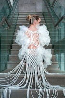 A person is seated on stairs, wearing an intricate, avant-garde outfit made of white fabric with net-like patterns and long flowing strands. The attire has pronounced puffed sleeves and a fitted back design. The person's hair is styled in an elegant updo, adorned with delicate chain decorations.
