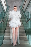 A person stands on a staircase wearing an elaborate white dress made of voluminous ruffles with long fringe elements extending from it. The dress is striking and avant-garde, paired with minimalist sandals. The setting is modern with glass railings and concrete steps, adding an industrial feel.