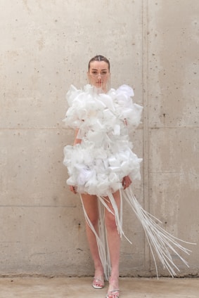 A person stands against a textured concrete wall wearing an avant-garde garment made of voluminous white tulle or fabric with long white strands extending from it. The outfit includes a facial decoration made of thin vertical lines. The setting appears minimalist and industrial.