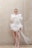 A person stands against a textured concrete wall wearing an avant-garde garment made of voluminous white tulle or fabric with long white strands extending from it. The outfit includes a facial decoration made of thin vertical lines. The setting appears minimalist and industrial.