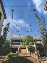 Traditional architecture with a two-story building featuring a tiled roof is surrounded by greenery under a clear blue sky. Wind chimes hang from wires creating a sense of serenity and movement.
