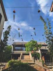 Traditional architecture with a two-story building featuring a tiled roof is surrounded by greenery under a clear blue sky. Wind chimes hang from wires creating a sense of serenity and movement.