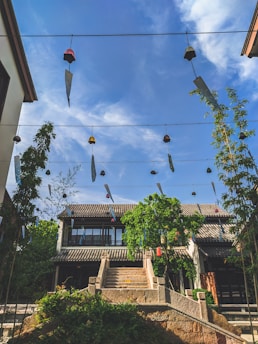 Traditional architecture with a two-story building featuring a tiled roof is surrounded by greenery under a clear blue sky. Wind chimes hang from wires creating a sense of serenity and movement.