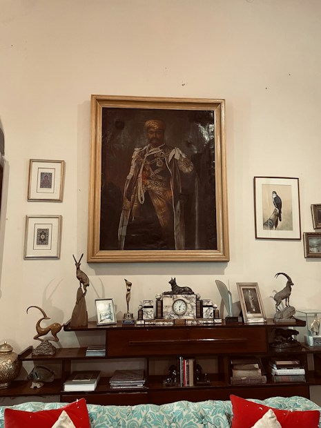 A warmly-lit living room features an elegant, traditional decor. A large framed portrait of a man in regal attire is prominently displayed on the wall. Surrounding the portrait are smaller framed artworks and photographs. The shelf beneath holds various decorative items, including statuettes of animals, a classic clock, and several books stacked orderly. Bright red cushions accent a blue and white patterned sofa, adding a pop of color to the room.