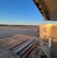 A large open runway with a clear blue sky in the background. In the foreground, the edge of a cargo area is visible with metallic components and a QR code on a metal panel. A small red vehicle is parked on the tarmac along with a few trolley-like structures.